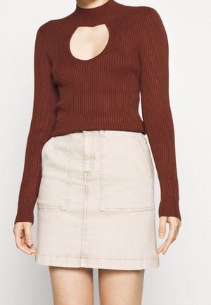 Žena nosící žebrovaný top s dlouhými rukávy v rezavé barvě s výstřihem na hrudi, doplněný béžovou krátkou sukni s vysokým pasem a kapsami.