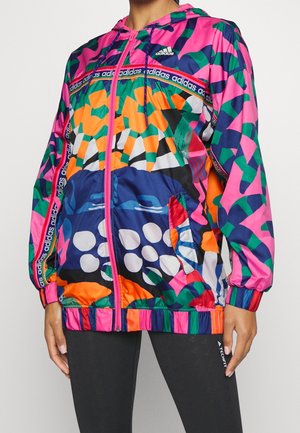 Person trägt eine bunte Adidas-Kapuzenjacke mit abstrakten Mustern in Pink, Grün, Orange, Blau und Weiß sowie schwarze Leggings.