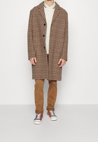 Rutig överrock i brunt och beige med slagkrage och knäppning framtill, över en gräddfärgad pikétröja, kombinerad med beige byxor och flerfärgade sneakers.