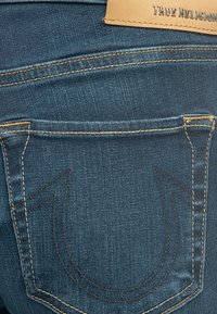 Poche arrière d'un jean en denim bleu avec des coutures jaunes, une broderie discrète en forme de fer à cheval, et une étiquette de marque en cuir tan indiquant "True Religion".