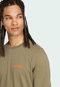 Oliven grøn T-shirt lavet af bomuld, med en rund hals og et orange "TERREX" logo på venstre brystområde.