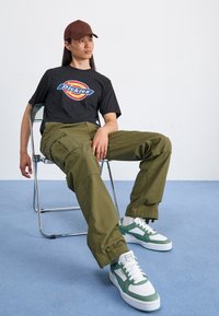 Černé tričko s grafikou "Pickles", olivově zelené kalhoty cargové a bílo-zelené tenisky. Sedí na kovové židli na světle modré podlaze.