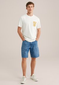 Bijela pamučna majica s žutim grafičkim uzorkom na lijevoj strani džepa, uparena sa plavim trapericama i bijelim sportskim tenisicama. Glatka tekstura, ležeran kroj.