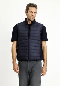 Granatowa, pikowana kamizelka z wysokim kołnierzem, zapinana na zamek, z kieszeniami po bokach. Gładka tekstura z poziomym przeszyciem.