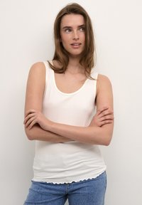Top in cotone bianco a coste con scollatura a barca e orlo scallop, abbinato a jeans azzurri chiari. Texture liscia e design aderente.