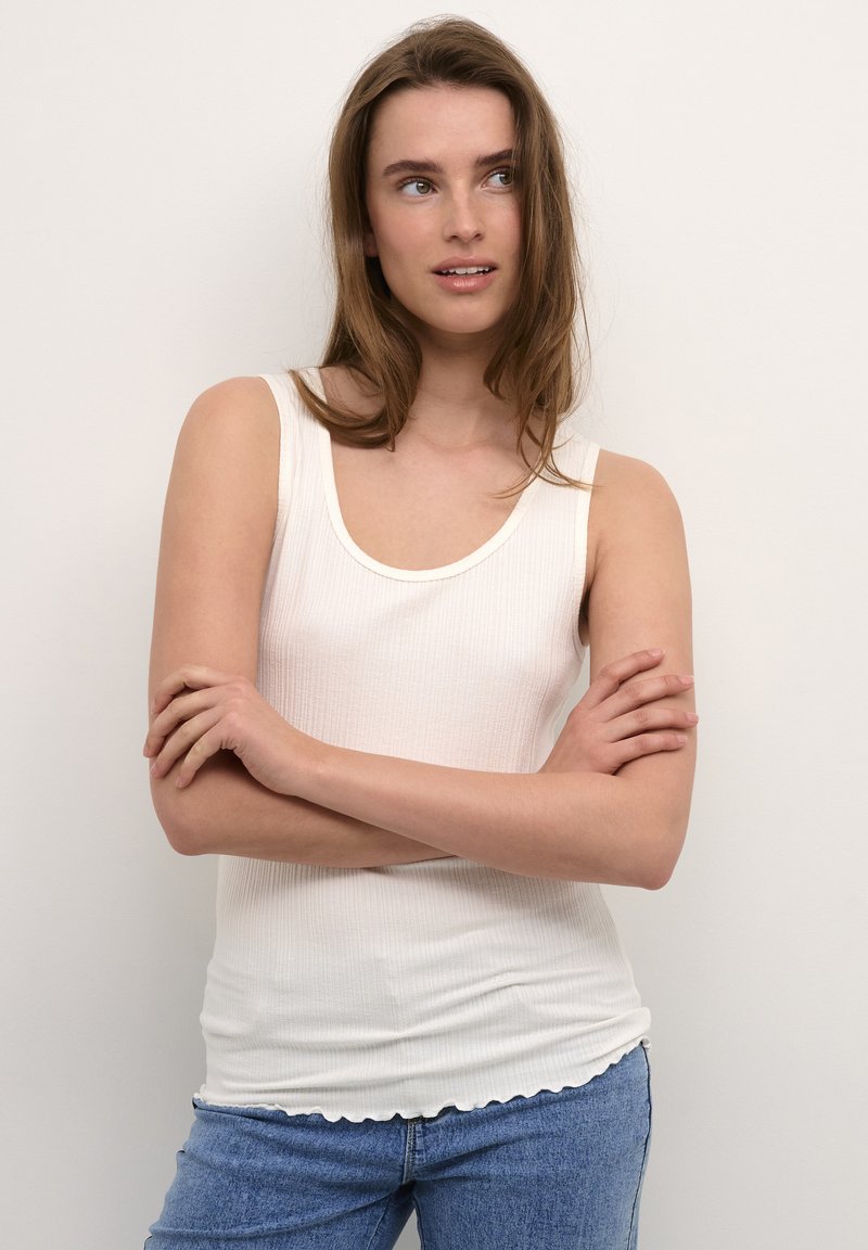 Valge soonikkoes tank top, millel on ümmargune kaelus ja laineline äär, koos helesiniste teksadega. Sile tekstuur ja keha lähedane disain.
