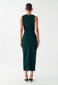 Robe midi sans manches verte avec une silhouette ajustée, fermeture éclair au dos et fente sur le côté. Tissu lisse avec un léger éclat ; longueur jusqu'à la cheville.