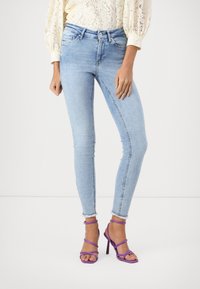 Jeans ajustados de color azul claro con dobladillo desflecado y diseño de cinco bolsillos, combinados con sandalias de tacón alto moradas y de tiras.