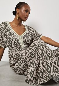 Maxi-jurk in een groen en beige ikatpatroon, met een diepe V-hals, korte mouwen en complexe kraalversiering langs de halslijn.