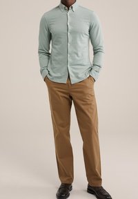 Man som bär en ljusgrön skjorta med knappar, bruna byxor och mörkbruna finskor, står med händerna i fickorna mot en neutral bakgrund.