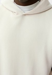 Sudadera con capucha de color beige claro hecha de una tela suave. Presenta un corte relajado, costuras visibles y un cuello redondo blanco superpuesto por debajo.