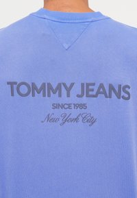 Sudadera azul claro con cuello redondo. Presenta un gran texto gris que dice "TOMMY JEANS" y texto más pequeño "Desde 1985 Nueva York" debajo.