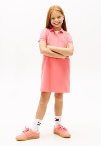 Χαμογελαστή κοπέλα με κόκκινα μαλλιά, που φοράει κοραλλί πόλο φόρεμα, λευκές κάλτσες crew με λογότυπα και κοραλλί αθλητικά παπούτσια, στέκεται με σταυρωμένα τα χέρια.