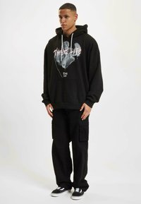 Sudadera negra de gran tamaño con un estampado gráfico que presenta "Thug Life", bolsillo frontal y cordones blancos, combinada con pantalones cargo negros sueltos y zapatillas.