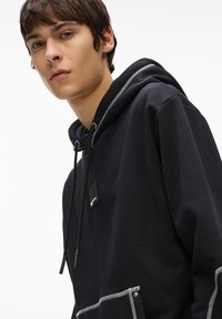 Melnā kapuci džemperis ar priekšējo kabatu, ar baltajiem šuvuma elementiem un mazā kvadrāta logotipu uz krūšu daļas. Gluda auduma tekstūra.