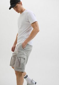 Helle graue Cargo-Shorts mit mehreren Taschen, kombiniert mit einem schlichten weißen T-Shirt und einer schwarzen Cap. Getragen mit weiß und schwarz gestreiften Socken und grauen Schuhen.