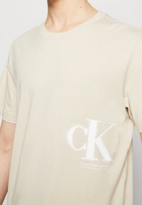 Camiseta de algodón beige con mangas cortas, escote redondo y logo y texto blancos "ck" impresos en la parte frontal. Textura suave.