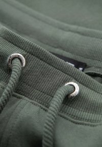 Tissu de hoodie vert olive avec texture côtelée, comportant des œillets en métal argenté et un cordon de serrage. Gros plan sur les coutures et les détails de design.