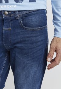 Vaqueros de mezclilla en un tono azul medio con corte recto, con un ligero desgastado, dos bolsillos frontales y cierre de botón metálico.