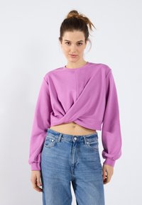 Lilla avkortet genser med lange ermer, med en vridd frontdesign. Kombinert med lyseblå denimjeans som har en avslappet passform.