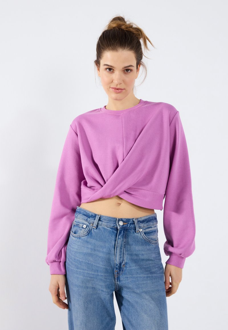 Lilla avkortet genser med lange ermer, med en vridd frontdesign. Kombinert med lyseblå denimjeans som har en avslappet passform.