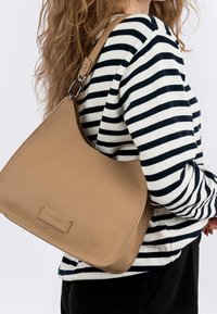 Vaalean beige-nahkainen olkalaukku, jossa on teksturoitu pinta ja hienovarainen logopatchi. Laukussa on leveä hihna ja kolmiomainen muoto.