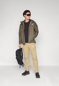 Jachetă impermeabilă de culoare verde măsliniu, tricou negru, pantaloni cargo bej, rucsac negru și adidași. Caracteristici includ o glugă și fermoare pe pantaloni.