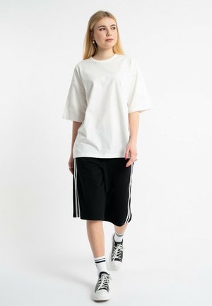 Młoda kobieta idzie, ubrana w luźny biały t-shirt, czarne krótkie spodenki do kolan z białymi paskami, paskowane skarpetki i czarne trampki, na białym tle.