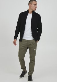 Schwarze Rippstrickjacke, weißes T-Shirt, olivgrüne Cargo-Hose mit Taschen Details und schwarze Sneaker. Einfaches, funktionales Outfit.