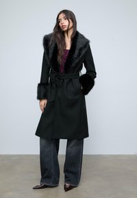 Donna che indossa un cappotto nero con cintura, colletto e polsini in pelliccia, jeans scuri a gamba larga e scarpe a punta su uno sfondo semplice.