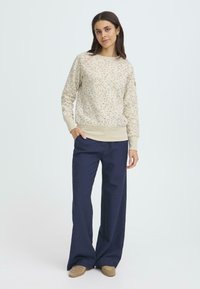 Felpa beige chiaro con macchie colorate, polsini e orlo a costine; abbinata a pantaloni navy a gamba larga e calzature tan.