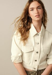 Veste en denim blanche coupée avec un col, des boutons-pression argentés et deux poches poitrine, dotée de manches retroussées et d'une texture lisse.