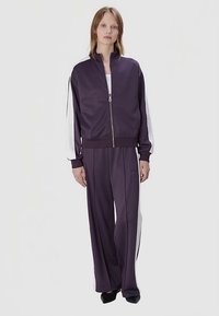 Fioletowy sportowy dres z białymi paskami po bokach. Tkanina ma gładką fakturę. Kurtka posiada zamek błyskawiczny z przodu i stojący kołnierz; spodnie mają szerokie nogawki.