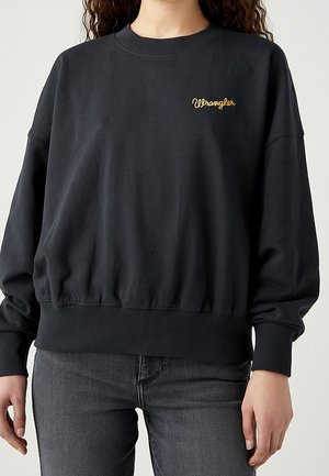 Person iført en sort Wrangler-sweatshirt med et lille gult logo på brystet og mørkegrå jeans mod en ensfarvet baggrund.