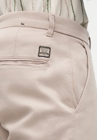 Jasnobeżowe spodnie z boczną kieszenią i małą czarno-białą metką GUESS przyszytą blisko pasa, noszone z wsuniętą w spodnie koszulą w paski.
