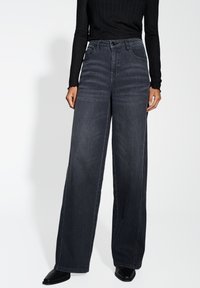 Jean en denim gris foncé à jambes larges et taille haute, avec cinq poches et des coutures contrastées. Porté avec un haut noir à manches longues côtelé.