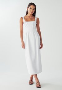 Žena v bezramienkových bielych midi šatách so štvorcovým výstrihom a čiernymi sandálmi s remienkami stojí proti svetlému pozadiu.