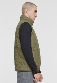 Olivgrüner gesteppter Weste mit hohem Kragen, Reißverschluss und seitlichen Taschen. Über einem schwarzen Langarmshirt getragen, im Profilansicht präsentiert.
