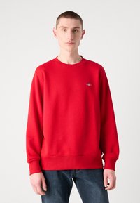 Rød sweatshirt med en struktureret overflade, rund hals og ribbede manchetter. Har et lille broderet logo på brystet.