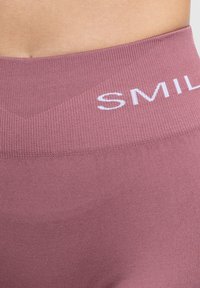 Leggings in tessuto elastico di colore rosa chiaro, con una ampia vita elastica che presenta la scritta "SMILE" in lettere bianche. Texture liscia e design aderente.
