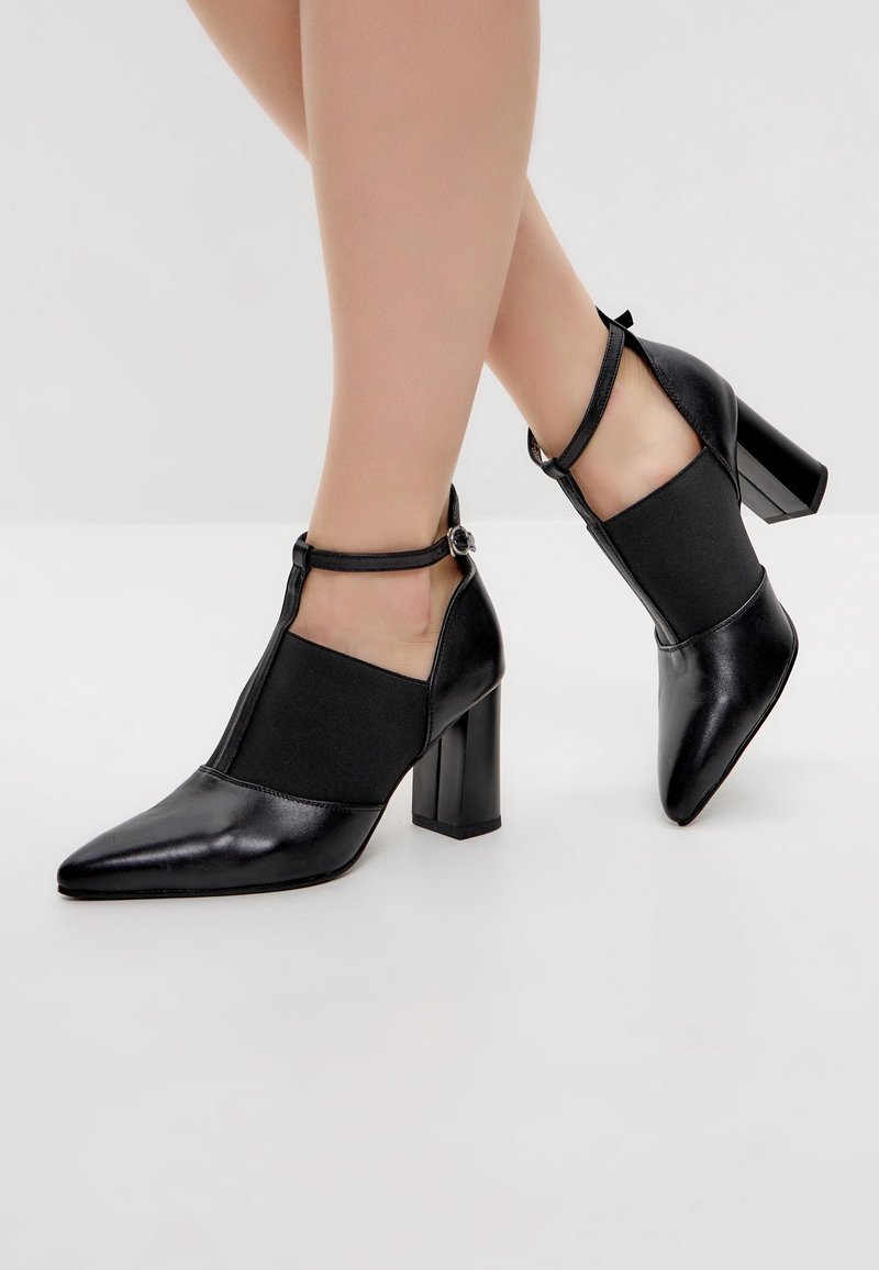 Chaussures en cuir noires à talons blocs avec brides en T et fermetures à boucle portées sur des jambes croisées sur un fond uni clair.