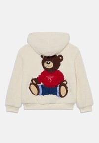 Kremno obarvan flis hoodie z velikim vezenim motivom medveda, ki nosi rdečo pulover in modre kratke hlače na zadnji strani. Zasnova s kapuco.