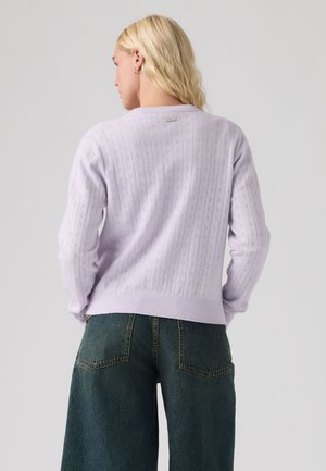 Femme blonde portant un pull en maille violet clair avec un motif ajouré et un jean large vert foncé, vue de dos.