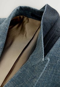 Interior de un blazer de denim azul que muestra un forro beige suave y un collar texturizado. Detalles de costura notables a lo largo de los bordes y la solapa.