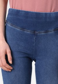 Leggings in denim blu con vita alta, superficie liscia e dettagli di cucitura visibili lungo le cuciture. Mano appoggiata accanto al tessuto.
