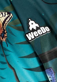 Tekstil med grønn-blå stof mønstrede med sommerfugle og blade, ledsaget af et hvidt og sort "WeeDo" mærke.
