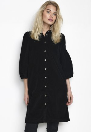 Robe chemise en velours côtelé noir avec un devant boutonné, des manches bouffantes trois quarts et une longueur au genou. Texture douce avec un design classique.