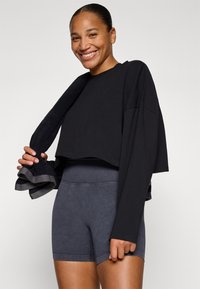 Zwart cropped lange mouwen top met brede mouwen, gecombineerd met donkergrijze high-waisted shorts. De persoon houdt een donkere handdoek vast.
