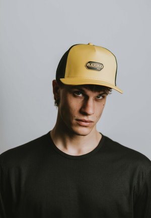 Young man wearing a yellow and black Blackskies Pro cap and a black ribbed shirt, looking directly ahead with a neutral expression.