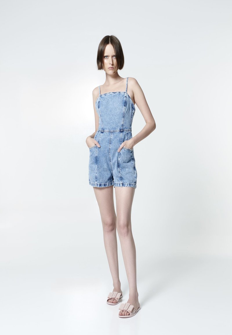 Salopette di jeans con spalline sottili, tasche frontali e vita aderente. Colore blu chiaro, texture sbiadita, pantaloni corti orlati. Abbinata a sandali beige.